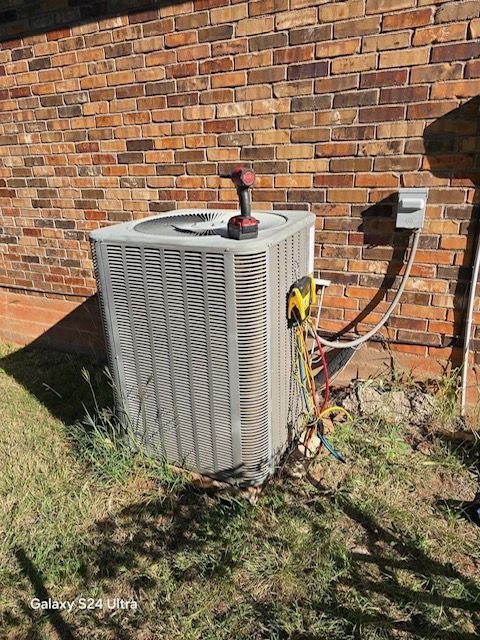 An air conditioning unit against a brick wall with tools resting on top and connected hoses, outdoors.