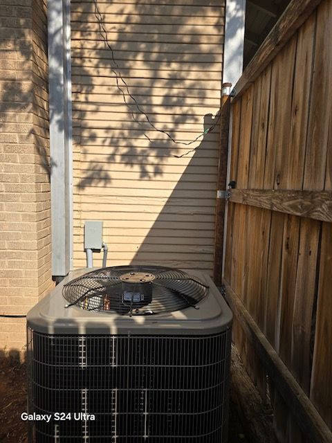 Outdoor air conditioning unit next to a wooden fence and siding under tree shadows.