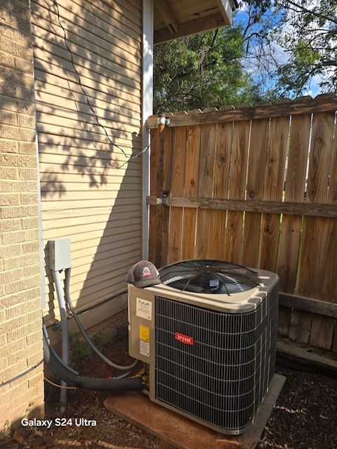 An air conditioning unit next to a wooden fence and building. A hat is resting on the unit.