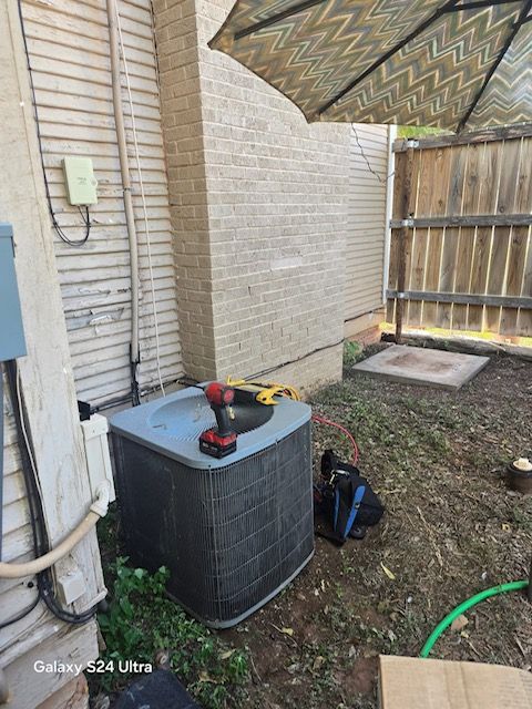 An air conditioning unit with a drill on top is in a yard next to a brick wall.