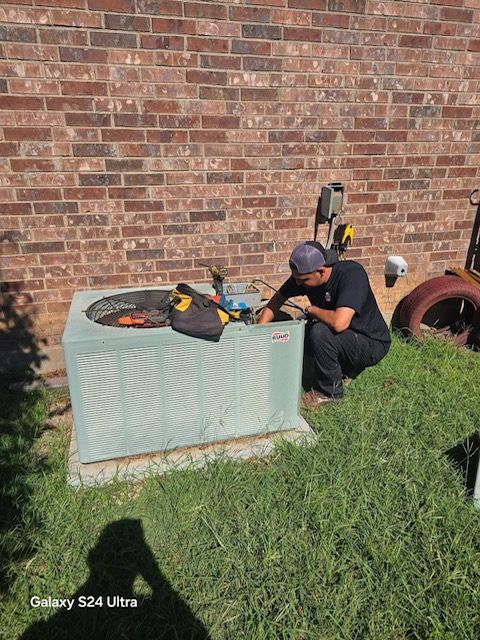 HVAC technician working on an AC unit near a brick wall outdoors.