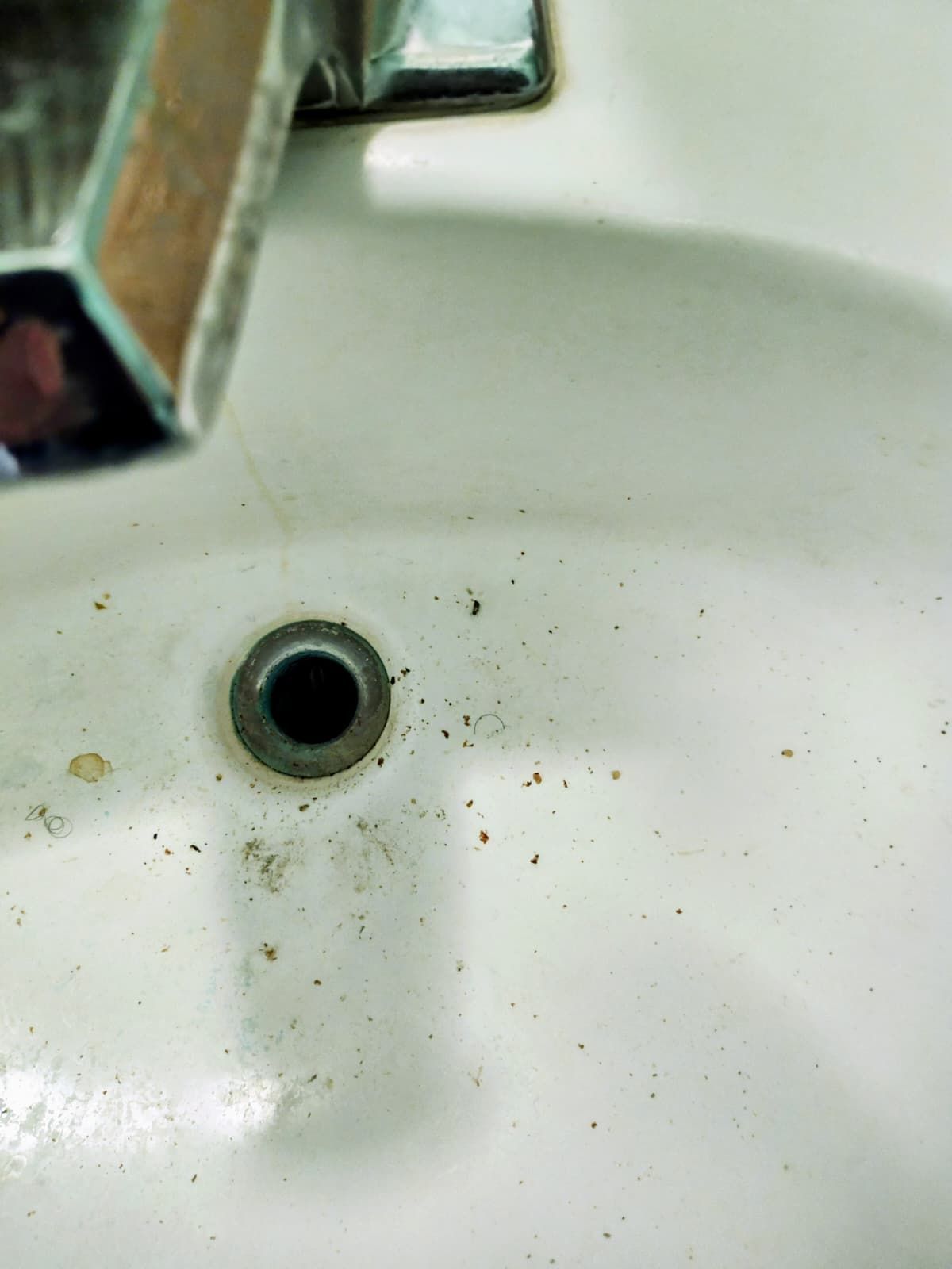 Close-up of a white bathroom sink drain with a chrome faucet at the top left and small dark specks around the basin