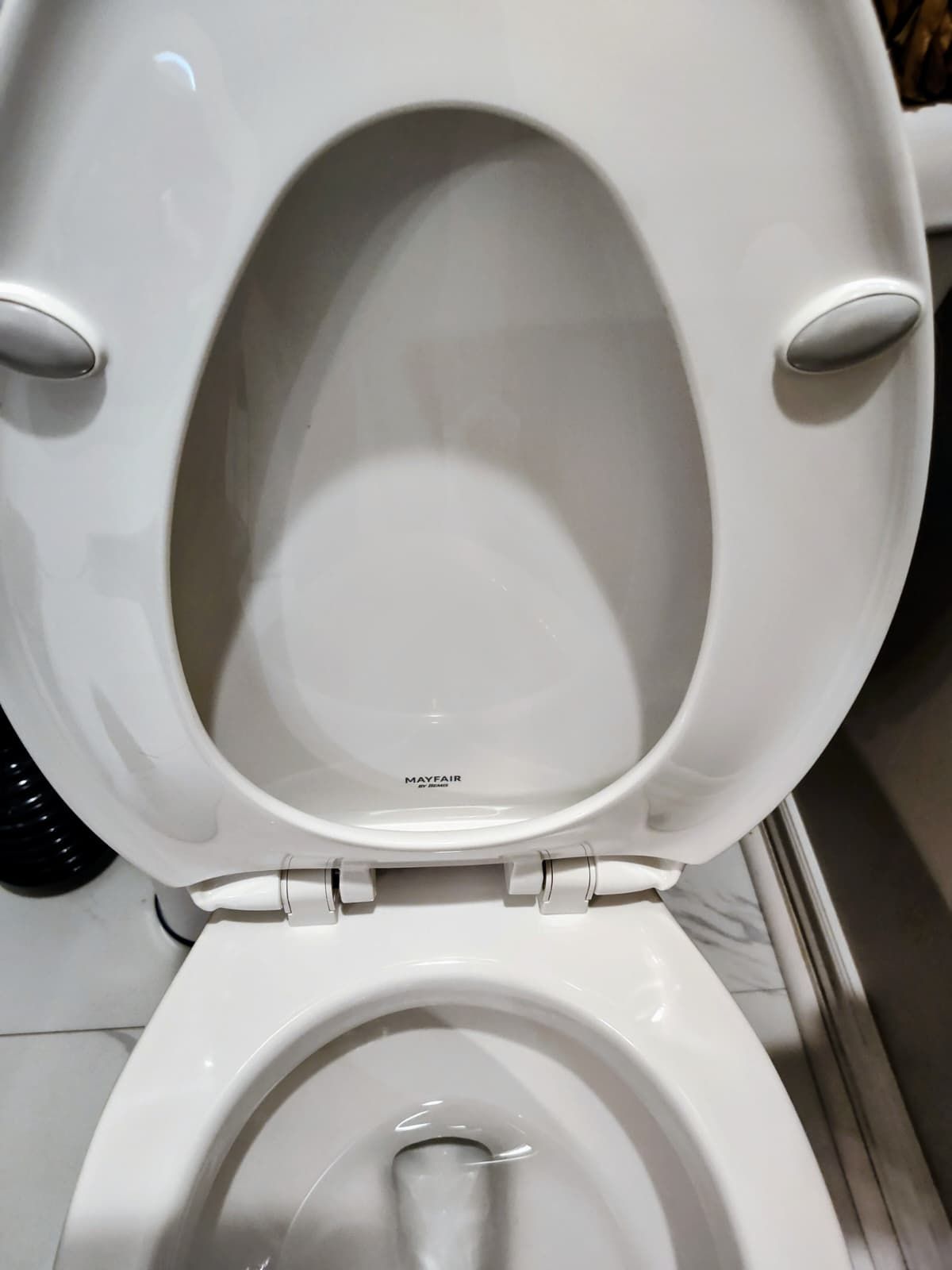 Open white toilet seat in a restroom, showing the bowl and hinge area.