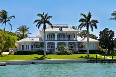 Large white waterfront mansion with palm trees and a clear blue sky, viewed from the water
