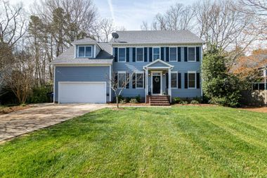 Two-story blue house with white trim, garage, and large front lawn in a wooded neighborhood.