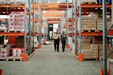 Two warehouse workers walking between stacked boxes and shelves in an industrial storage aisle.