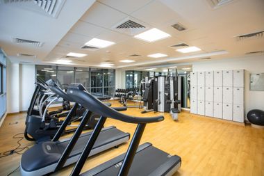 Modern gym with treadmills, exercise machines, mirrors, and lockers on a light wood floor.