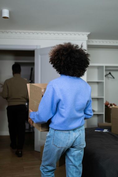 Person in a blue sweater carrying a box in a bedroom while another person walks away.