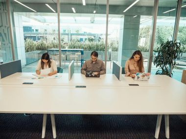 Three people seated at a long white table in a bright glass conference room, reading documents.