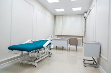 Empty medical exam room with teal treatment table, desk, chair, and bright white walls