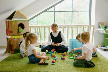Woman and three children playing with colorful blocks on a green carpet in a bright playroom