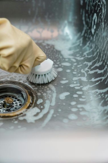 Gloved hand scrubbing a dirty kitchen sink with soap suds and a brush