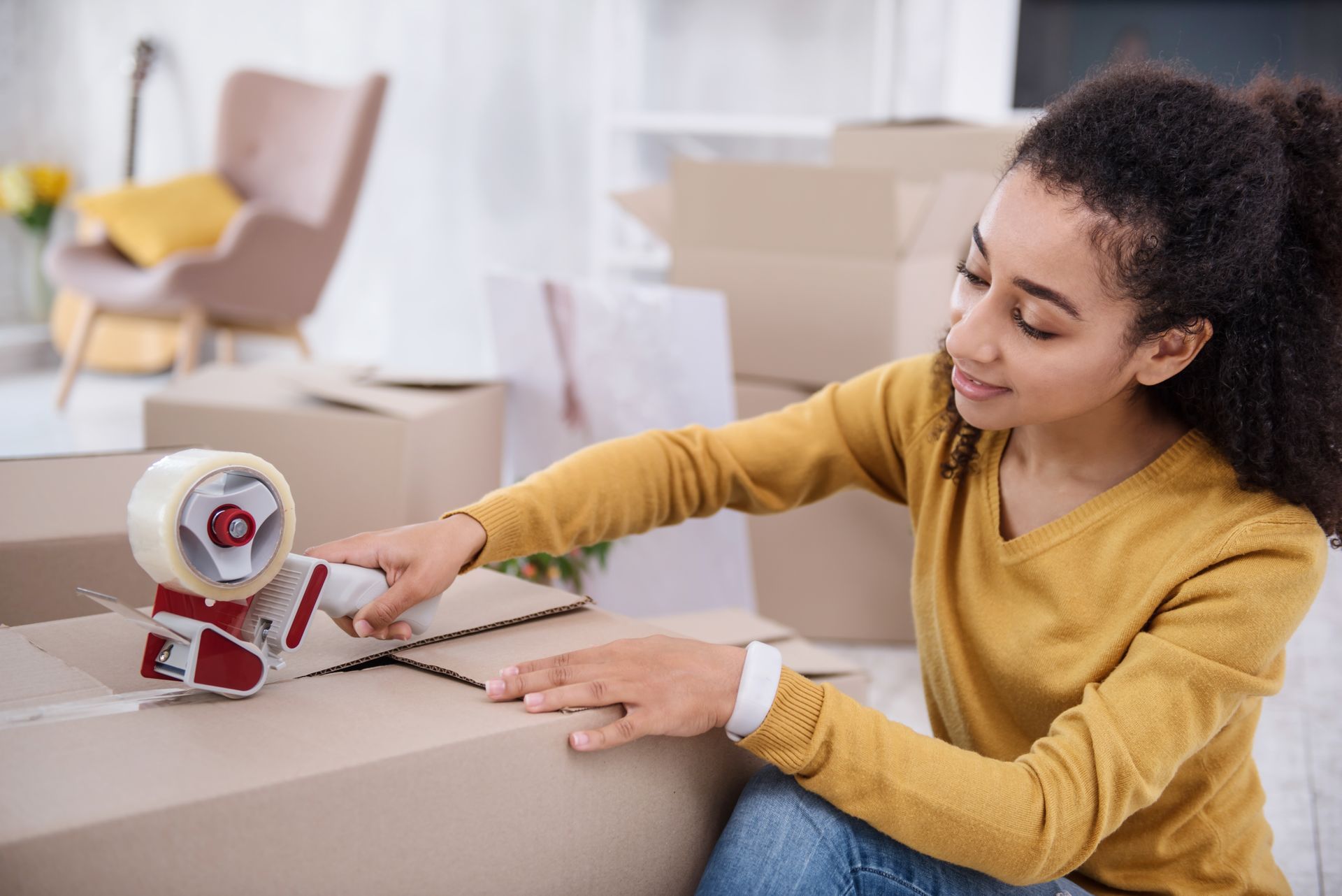 A woman is sitting on the floor using a tape dispenser to tape a box.