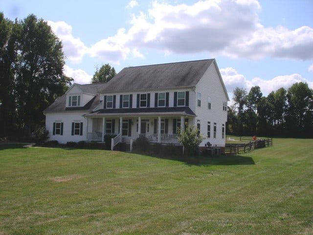White farmhouse with porch and dormer windows on a large grassy lawn under a partly cloudy sky