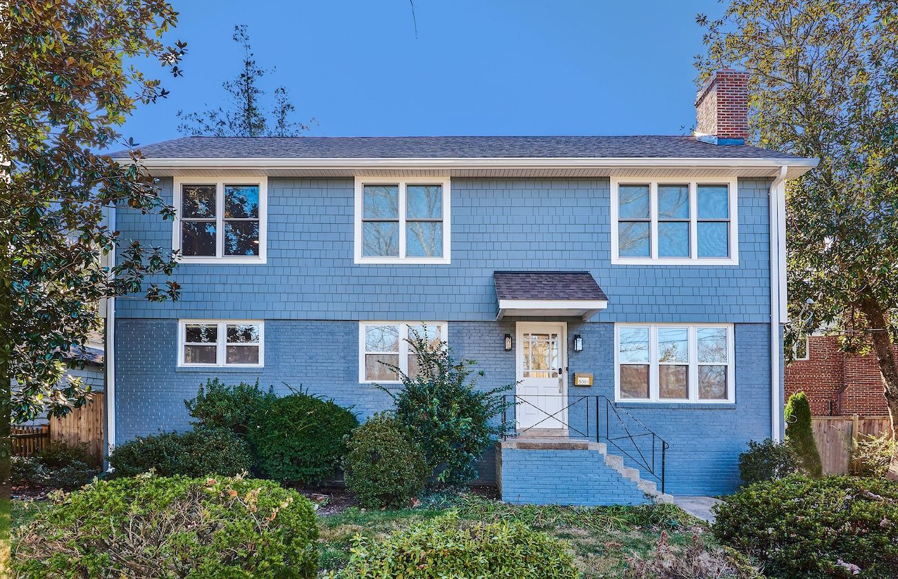 Blue two-story house with white trim and a front porch, surrounded by trees and bushes