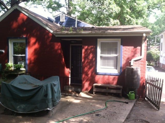 Small red house with white-trimmed windows, a covered grill, and a fence in a leafy yard