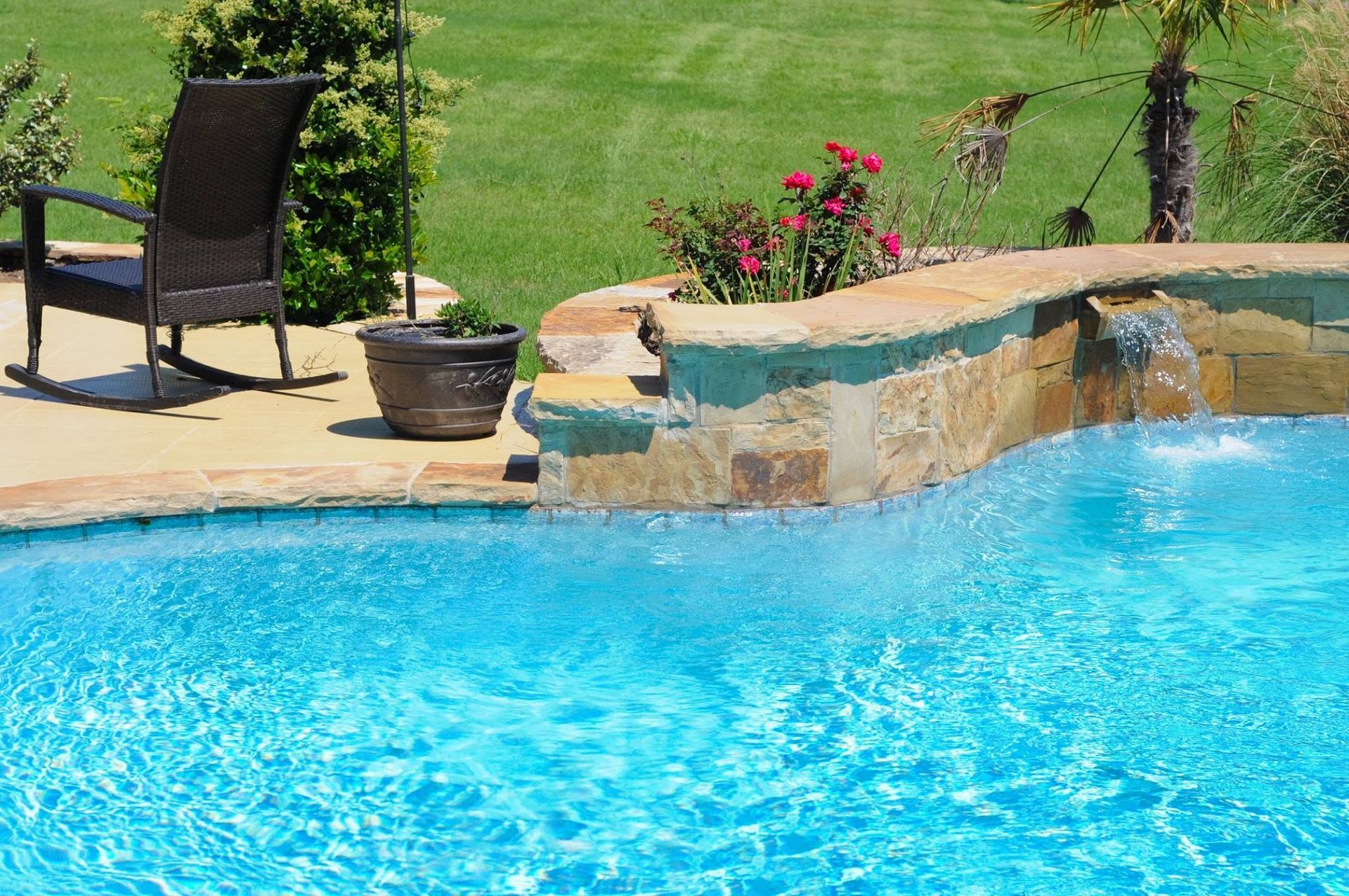 Backyard swimming pool with bright blue water, stone ledge, patio chair, and potted plant on the deck