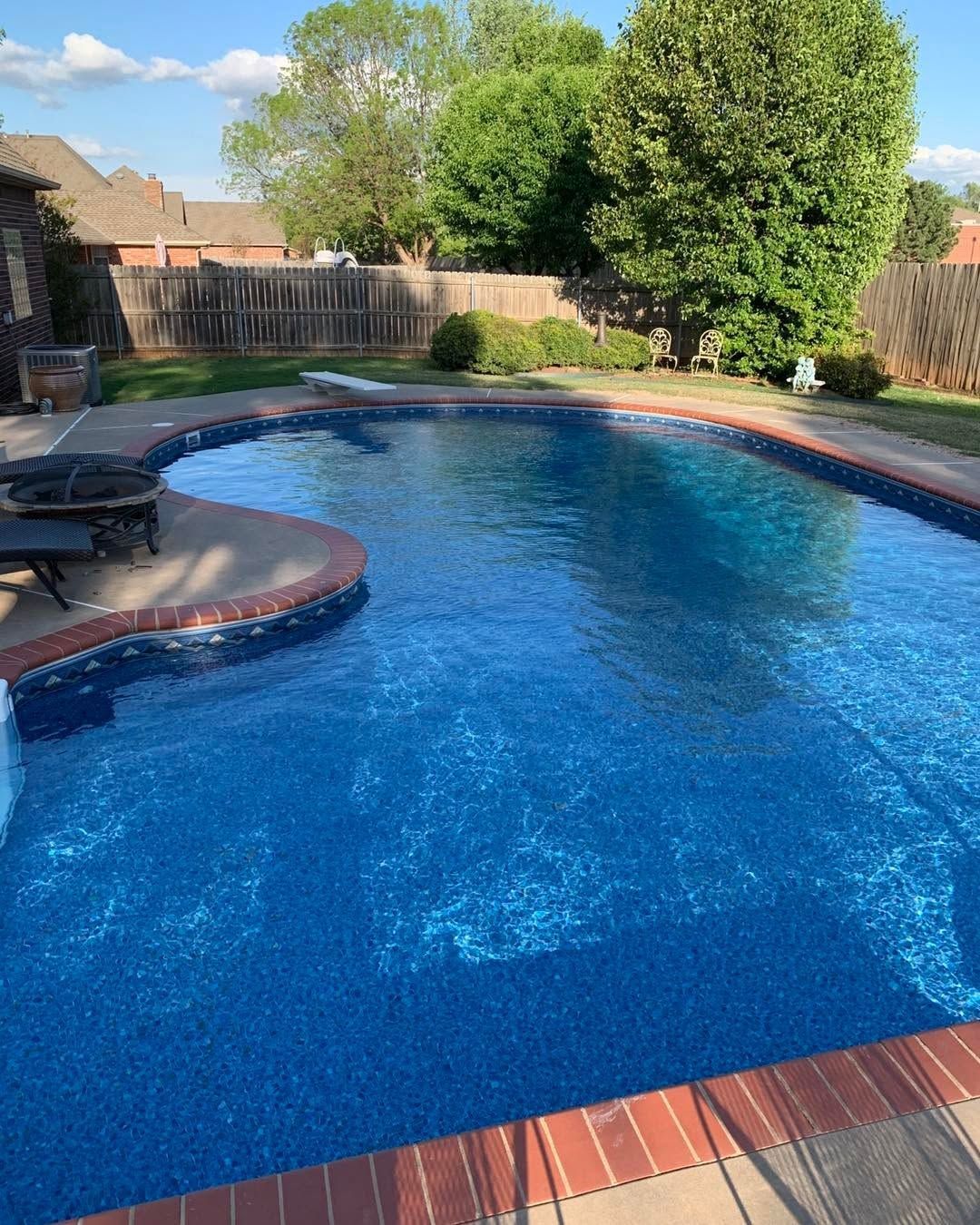 Backyard swimming pool with blue water, red brick coping, patio chairs, and trees in bright sunlight