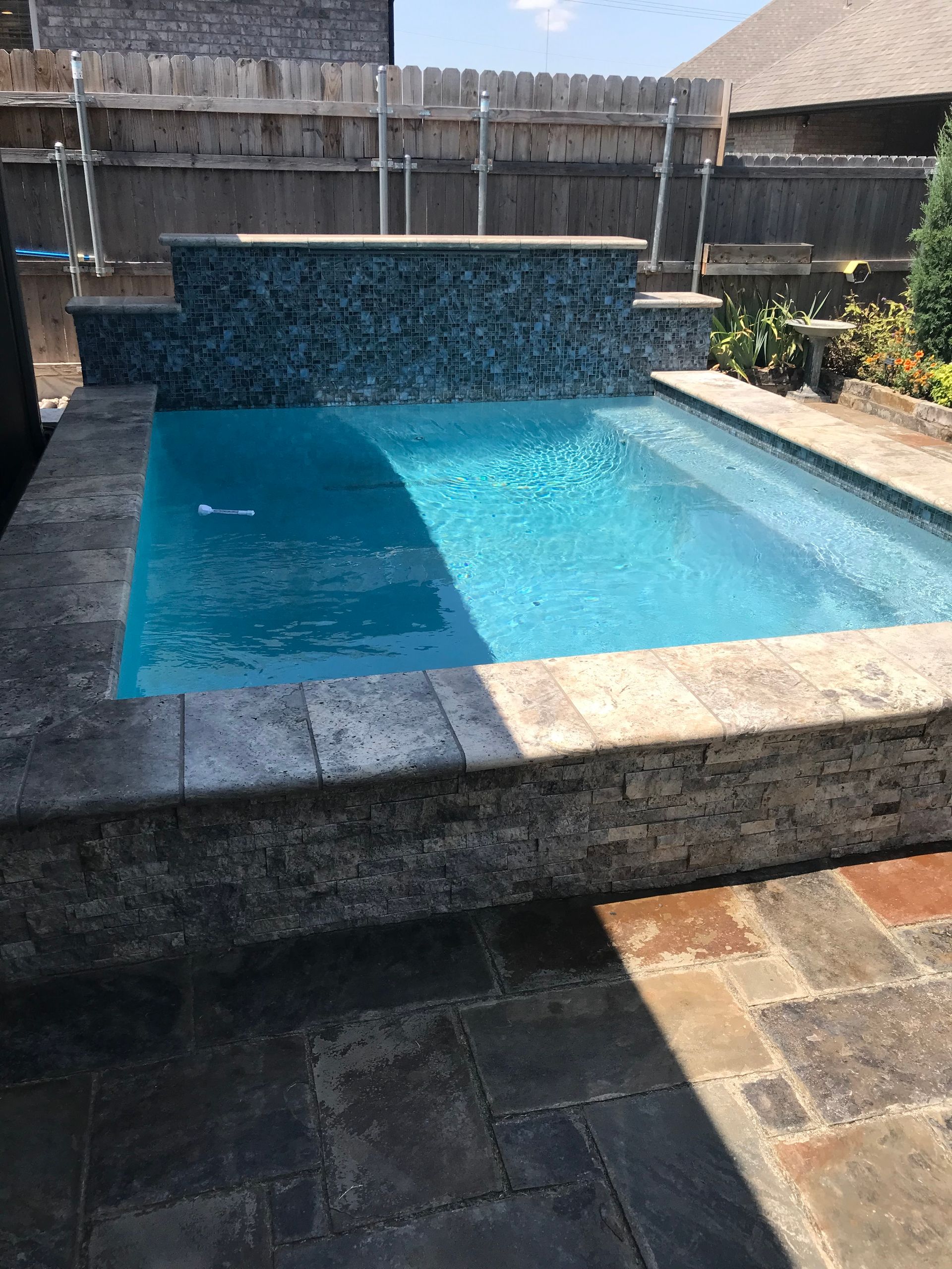 Small turquoise backyard pool with stone coping and a waterfall feature under bright sunlight