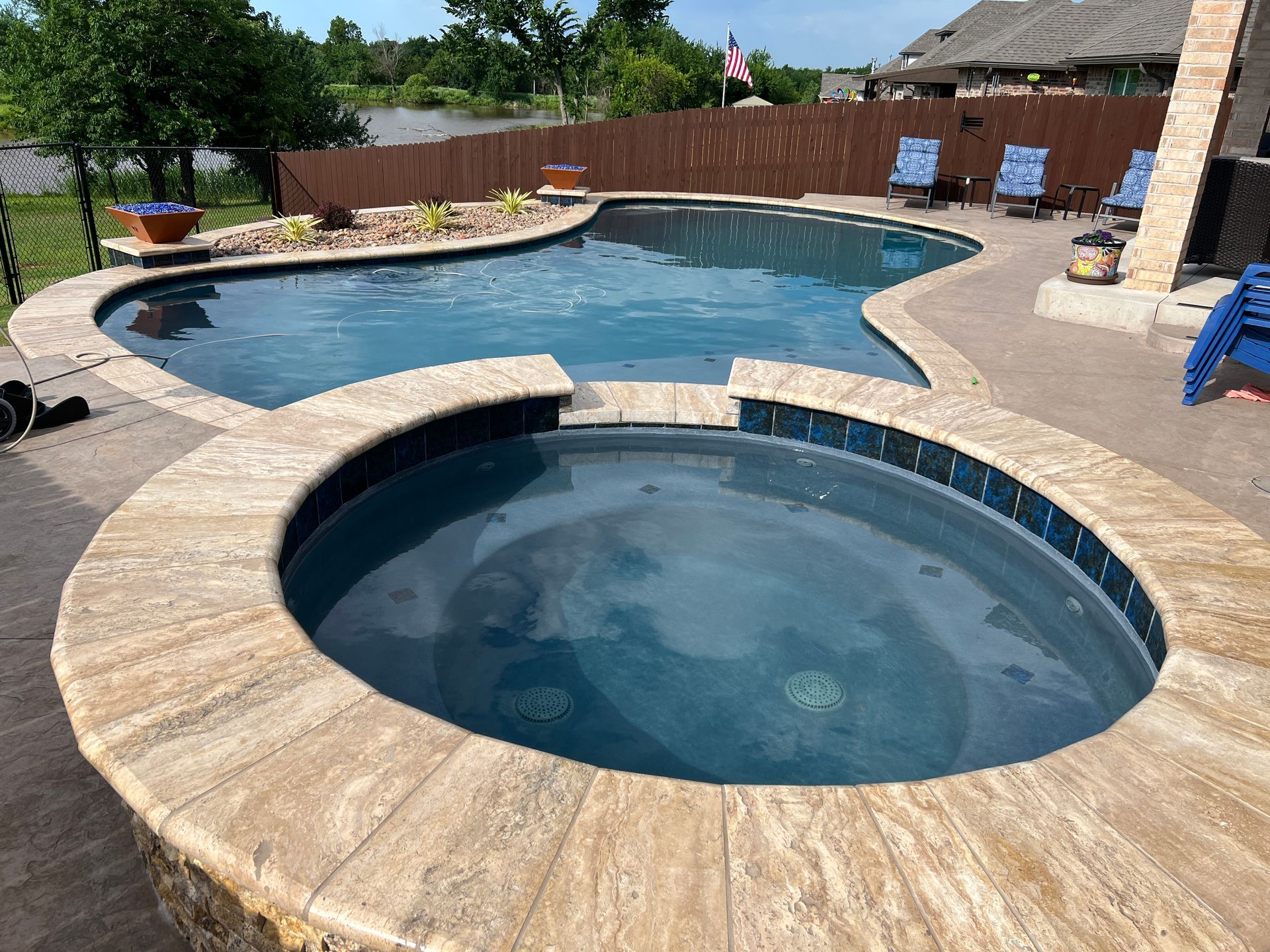 Backyard pool and hot tub with tan stone decking under a sunny sky