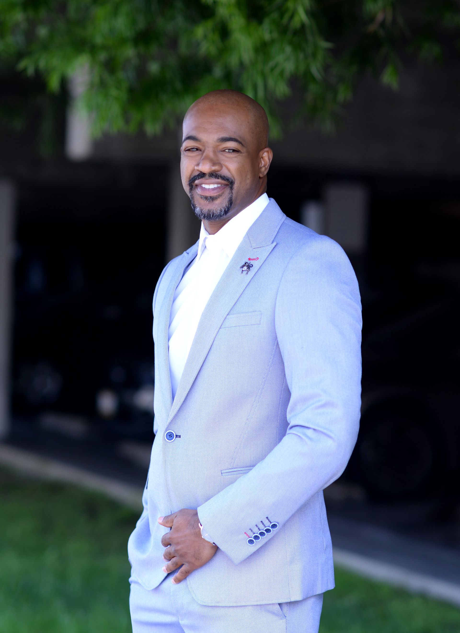 A man in a light blue suit is standing in front of a tree.