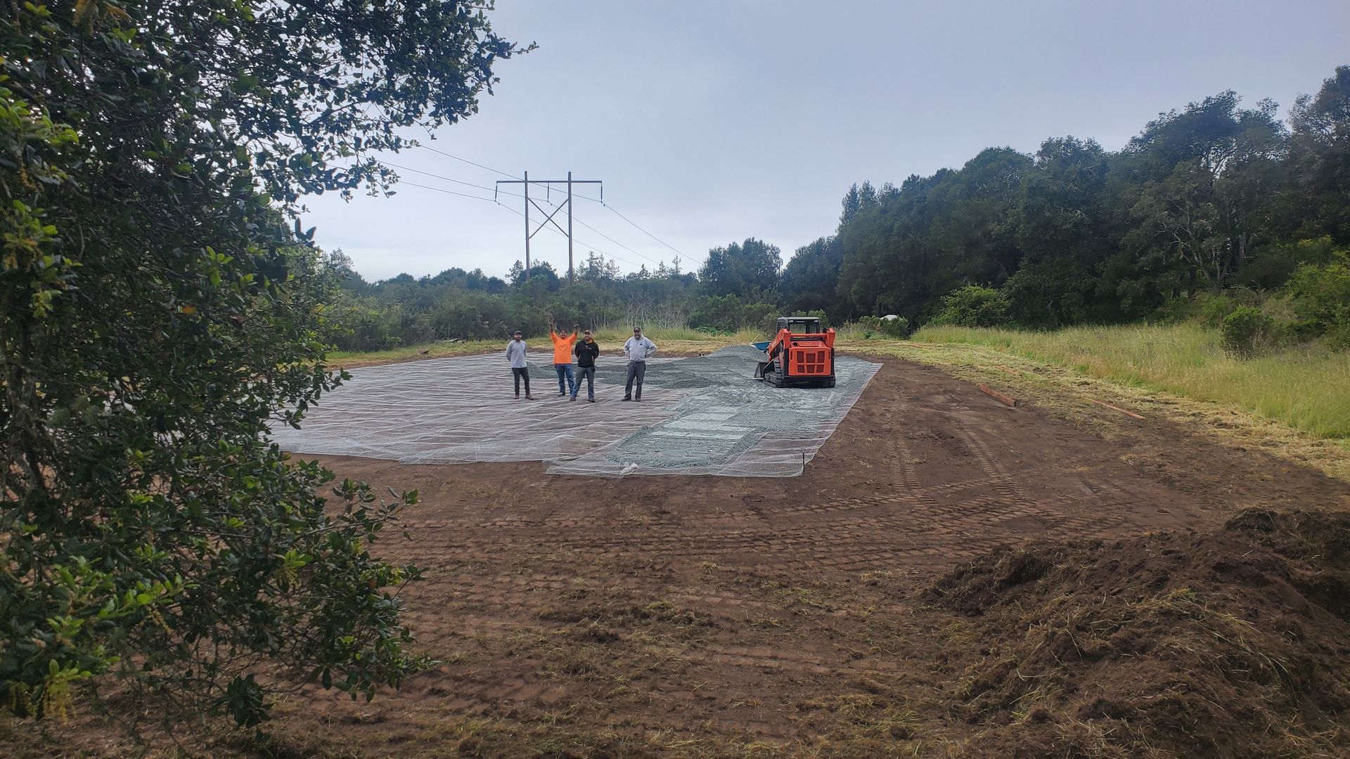 A construction site with people standing on a paved surface. A small tractor and power lines are in the background.