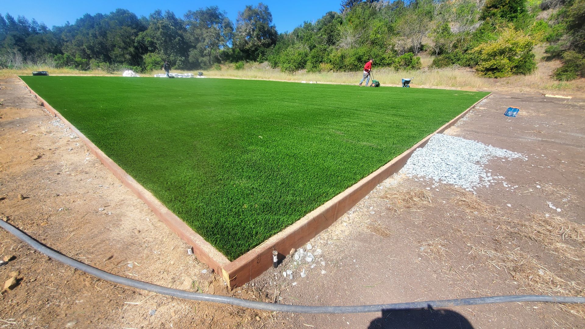 Artificial turf installation in progress; green turf within a wooden frame on dirt, with a worker in the distance.