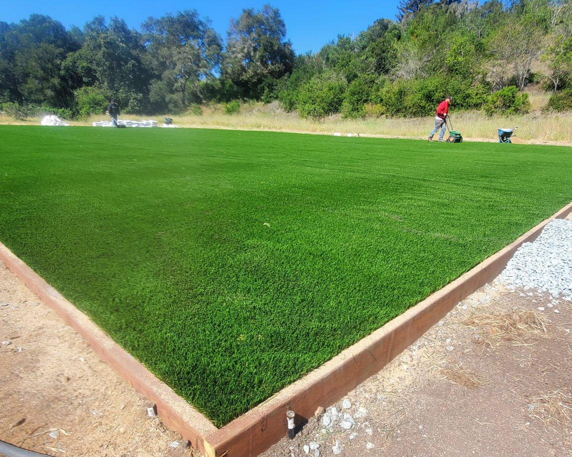 A patch of vibrant green artificial turf with a wooden border, workers in the background, and a sunny sky.