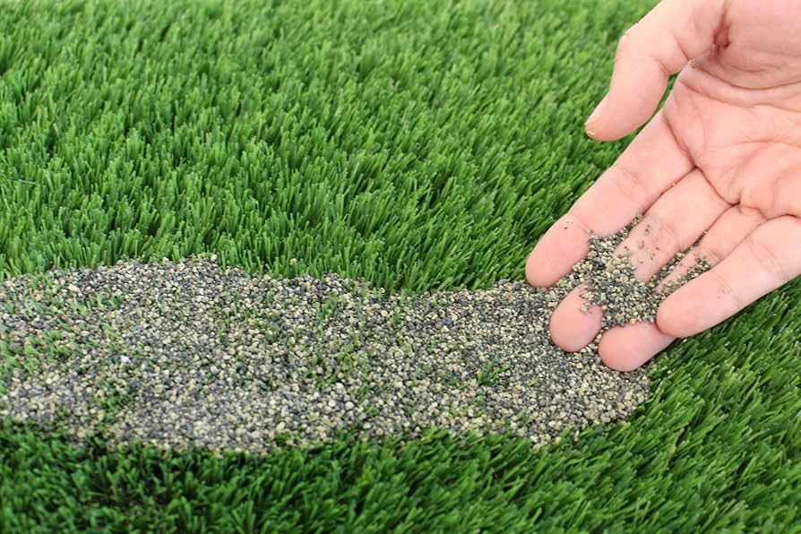 Hand sprinkles black granules onto a green artificial turf field, along a white line.
