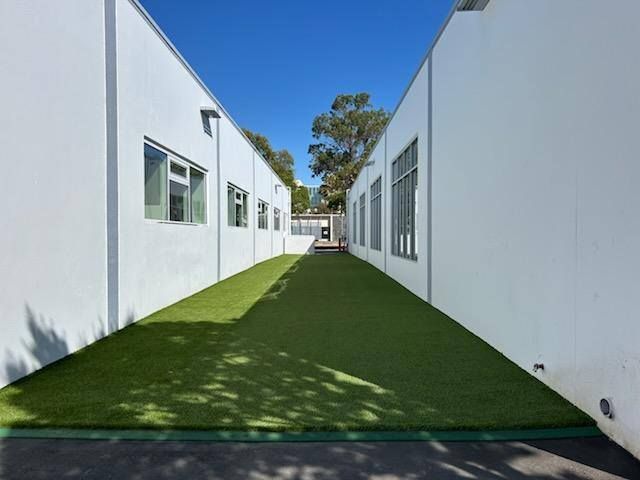 Narrow walkway between two white buildings with artificial grass; sunny day.