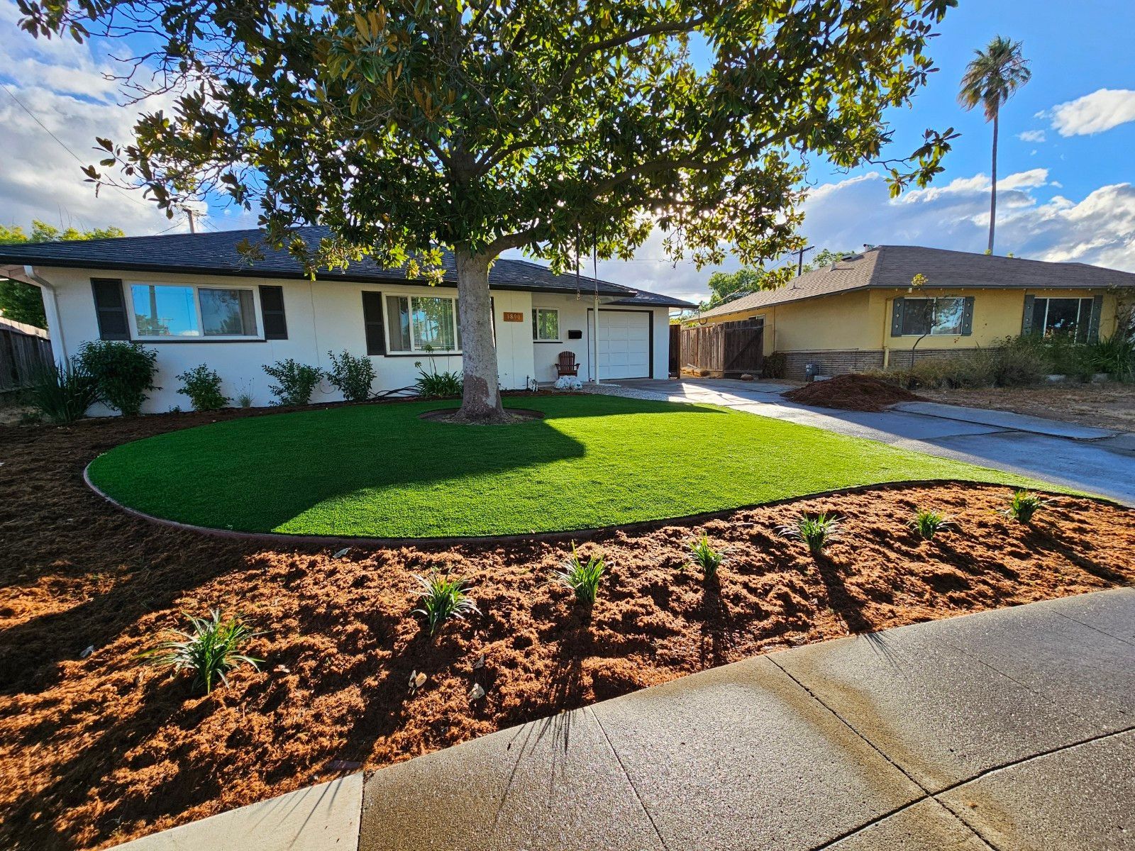 Green artificial lawn in a backyard, bordered by trees, a wooden fence, and patio seating.