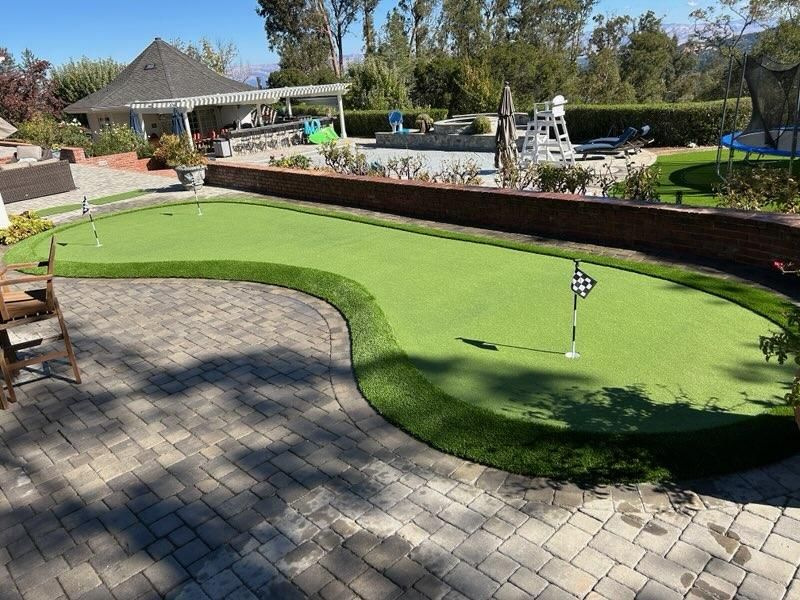 A backyard putting green with a flag. Brick patio and pool in background.