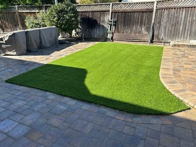 Green artificial turf on brick patio in backyard. Wooden fence and covered furniture in background.