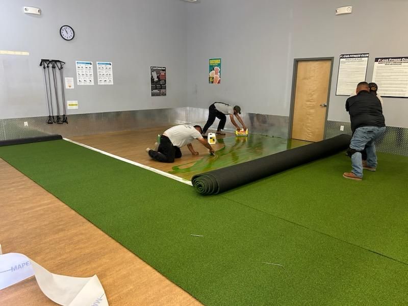 Three people installing green turf flooring in a gym. One rolls it out, others clean.