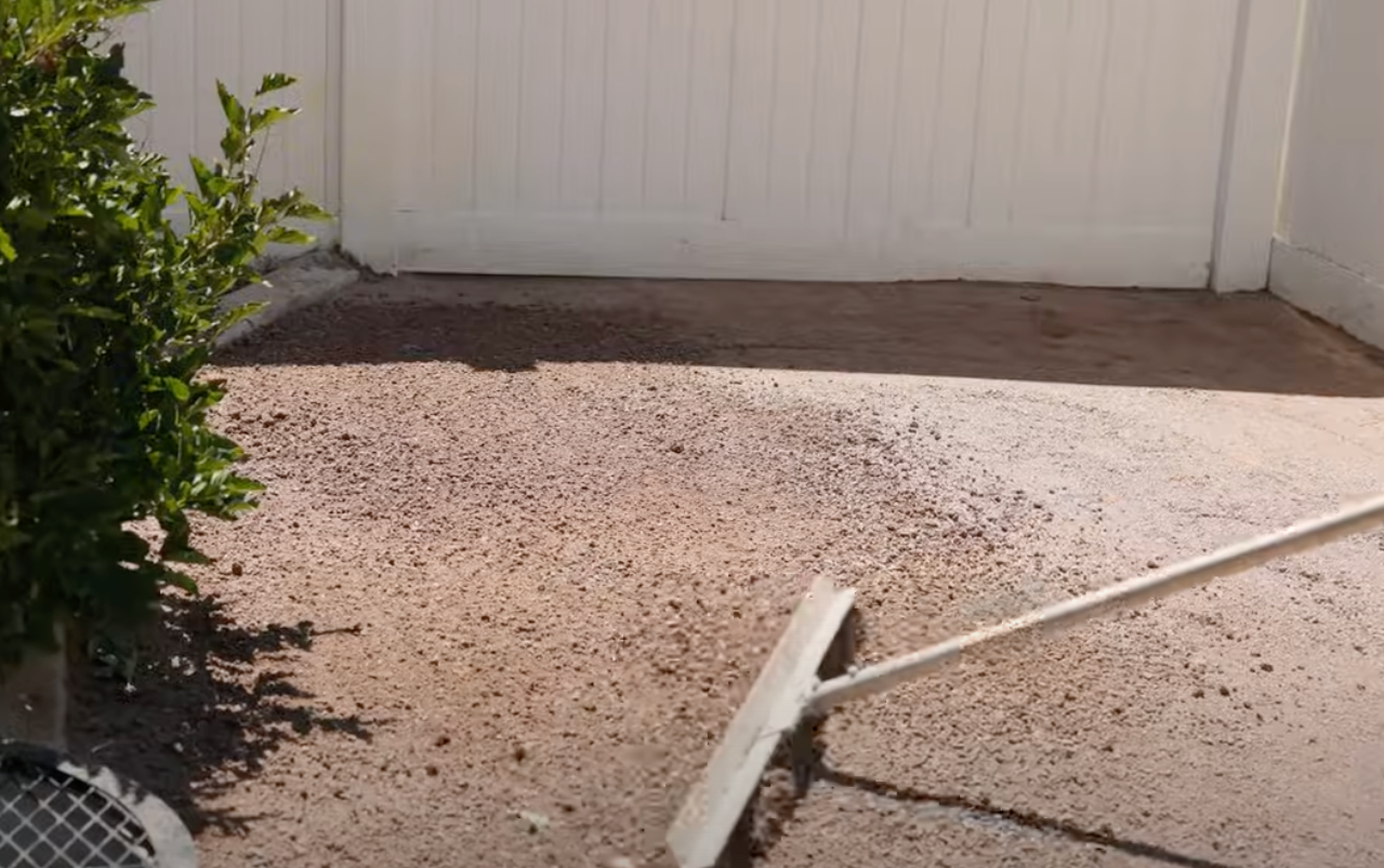 Raking gravel in a backyard; white fence in background; green bush on the left.