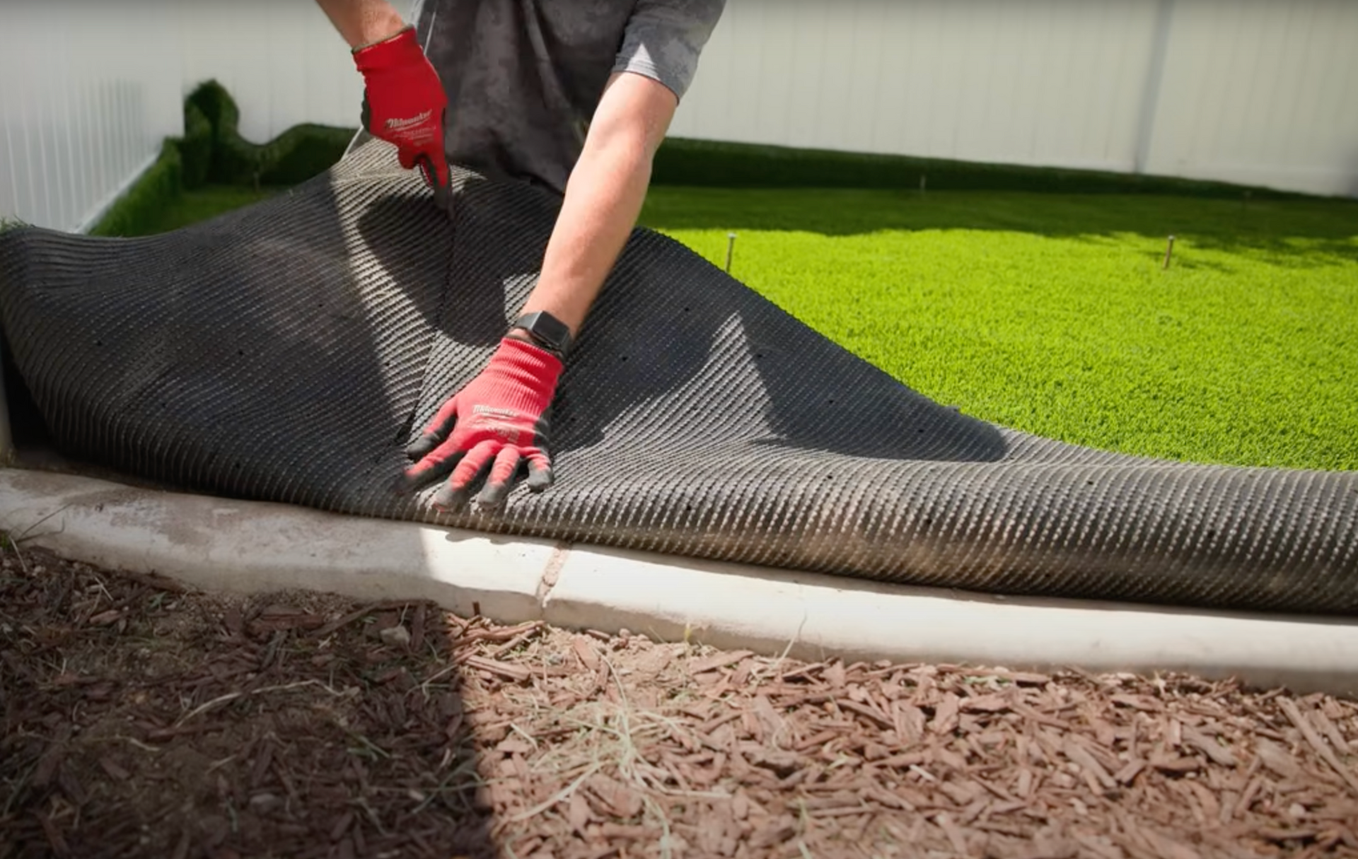 Person in red gloves unrolls artificial turf onto a concrete border, green lawn visible.