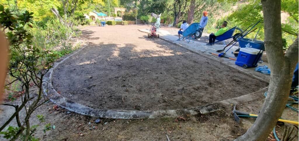 Workers prepare an oval garden bed, using wheelbarrows and tools in a sunny outdoor setting.