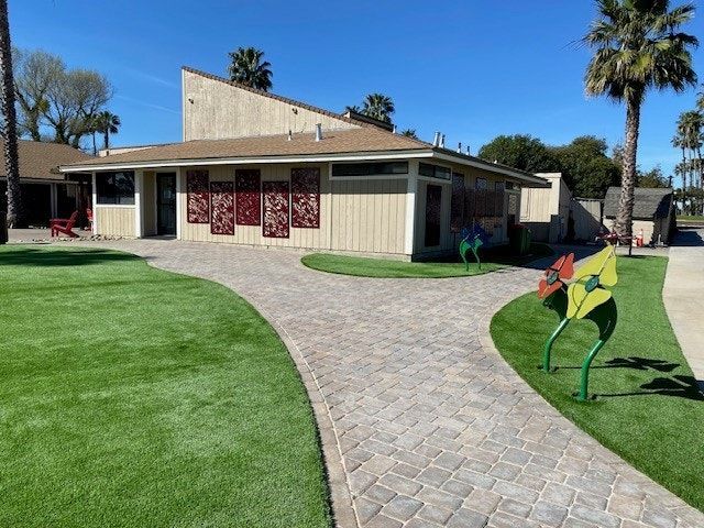 A beige building with a brick path and artificial green lawn.  Palm trees in the background under a blue sky.
