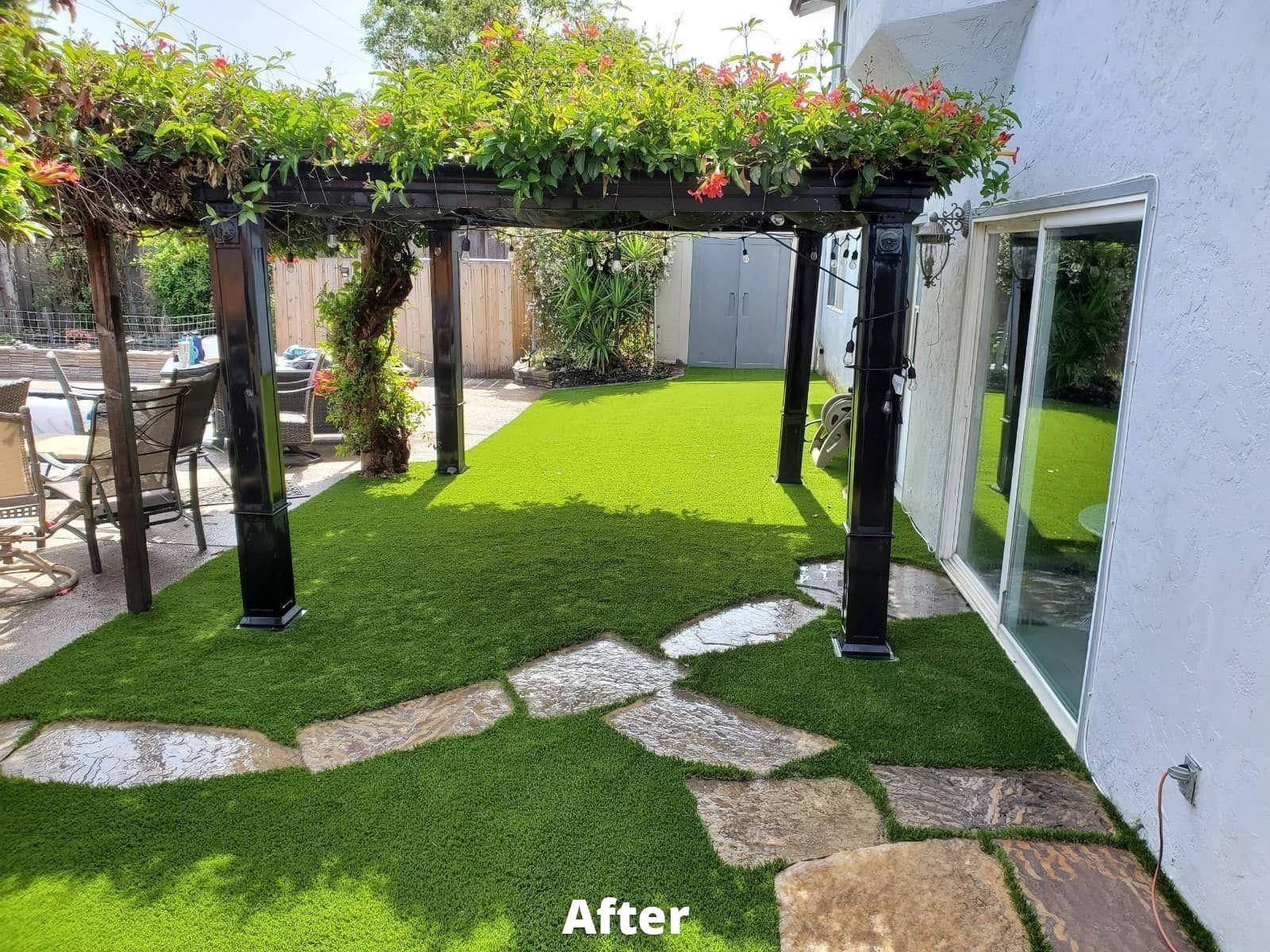 Backyard with a pergola covered in flowers, over a patch of green turf. A stone path leads to the area.