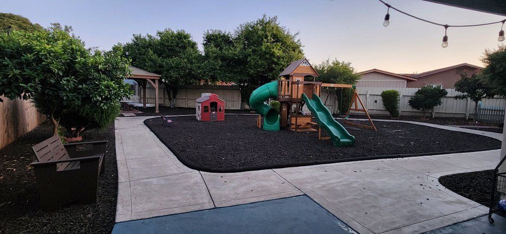 Backyard with playground equipment, benches, and trees. The ground is covered with black mulch.