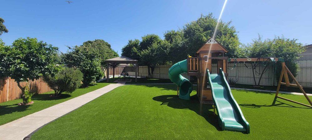 Backyard playground with a slide, swings, and a gazebo. Bright green grass and trees under a blue sky.