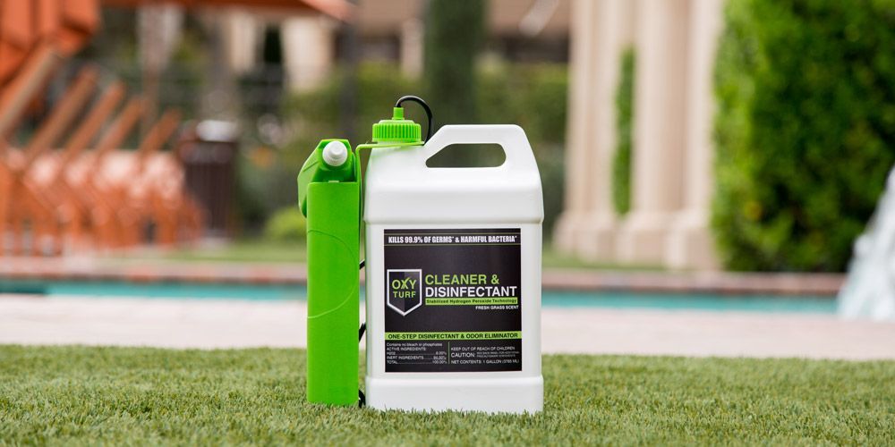 White and green disinfectant container next to a green sprayer, on green artificial turf. Background shows pool and trees.