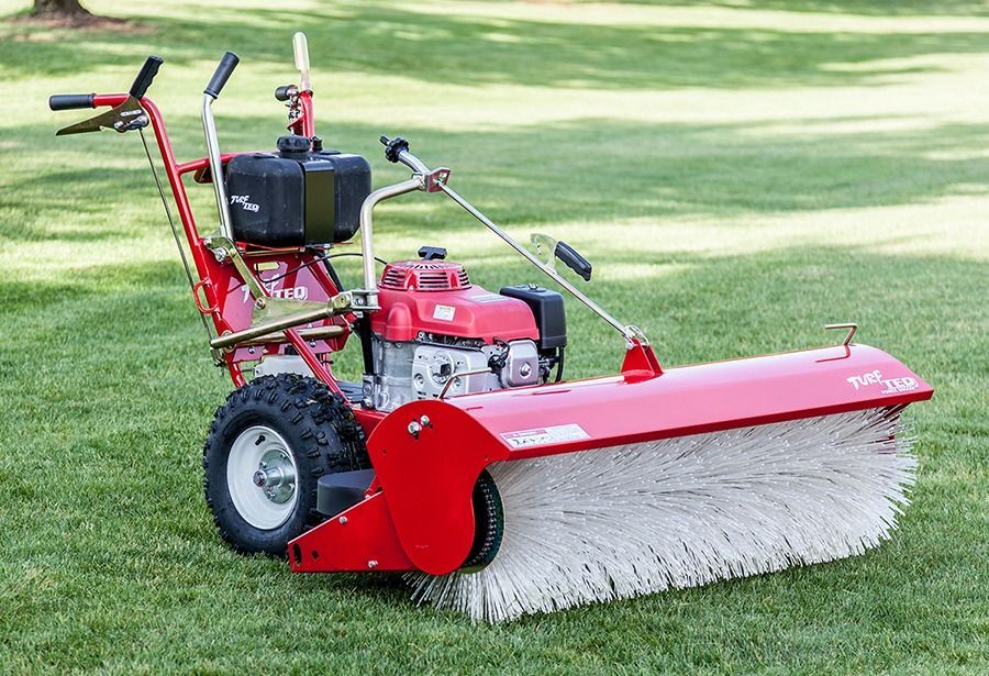 Red and black power broom on grass, used for sweeping, has a gas tank and motor.