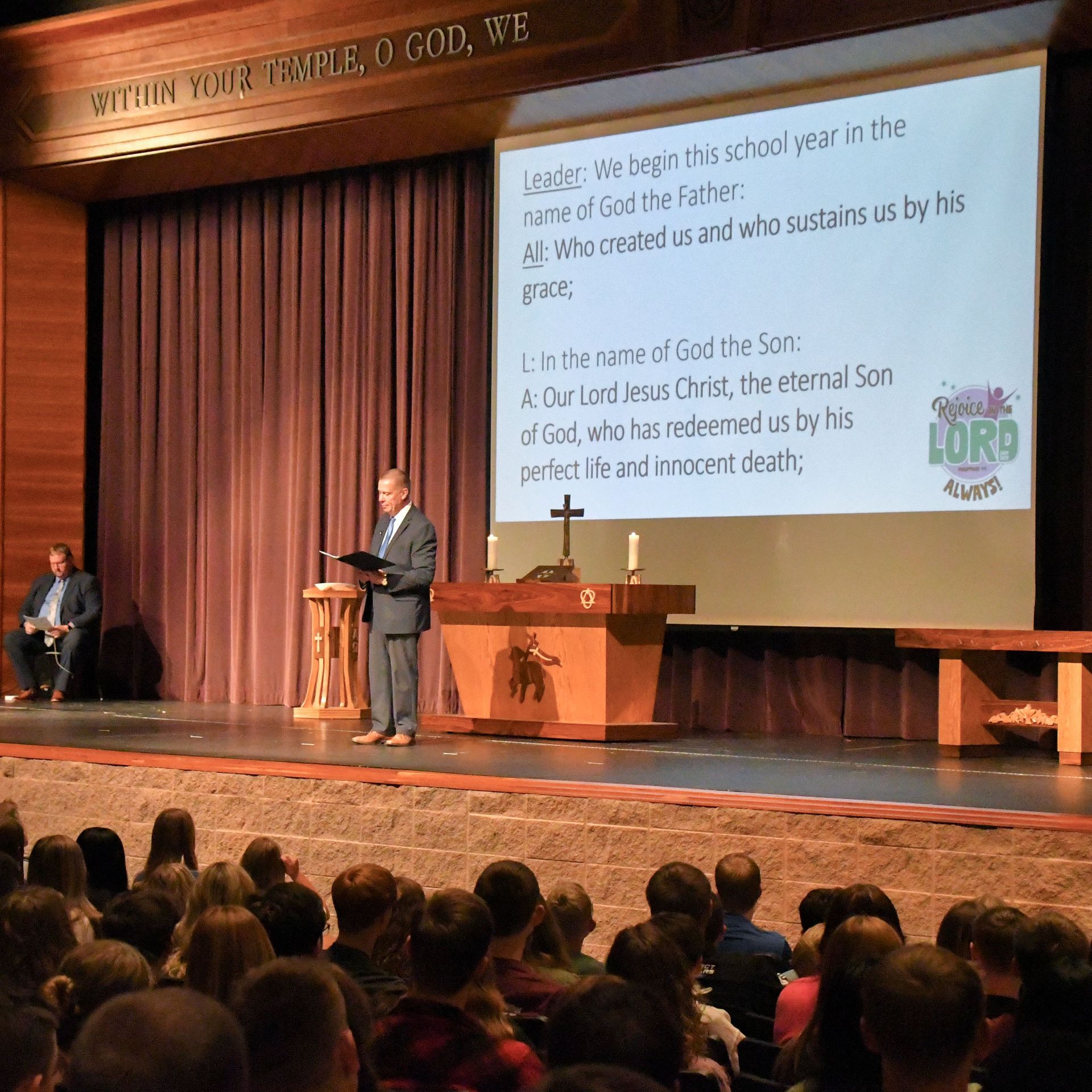Rev. Wenzel on the stage in the auditorium with the screen behind him and students in seats