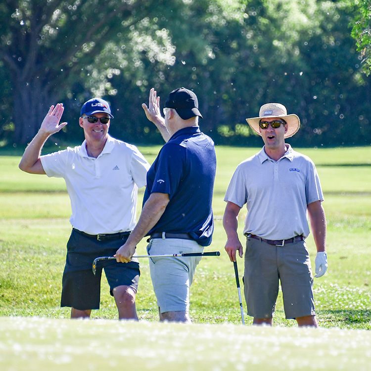 Two men giving each other a high 5 on the golf course while another man stands by, looking surprised