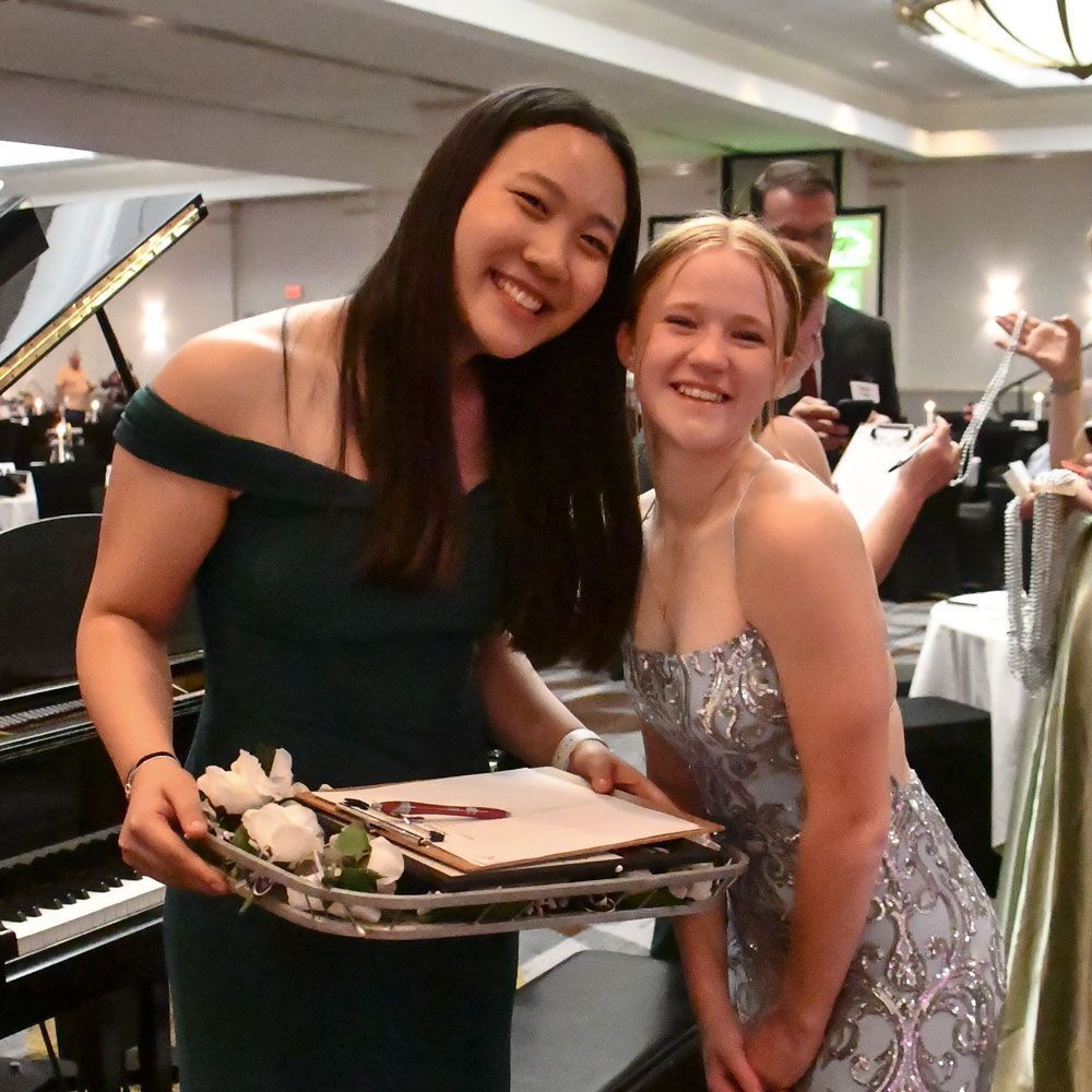 Two smiling high school girls in formal dresses, and holding a tray of roses for the Rose Lapel event