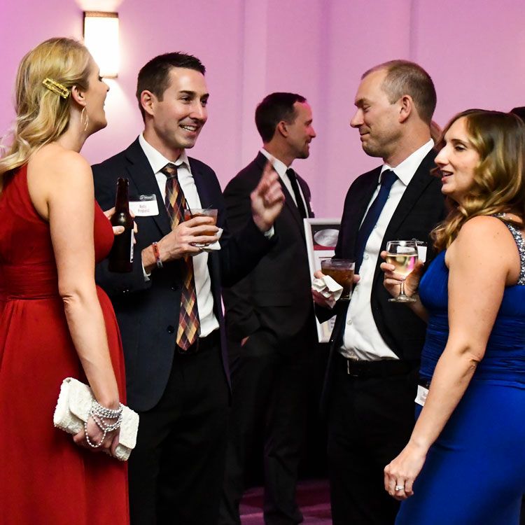 Two men and two women standing in a group. They are in evening attire, enjoying conversation and drinks