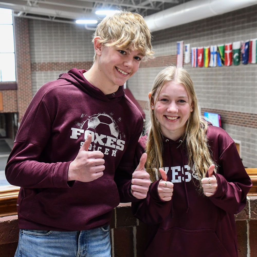 Happy boy and girl students with their thumbs up and the FVL Commons behind them. The photo was taken in the balcony area above and overlooking Commons
