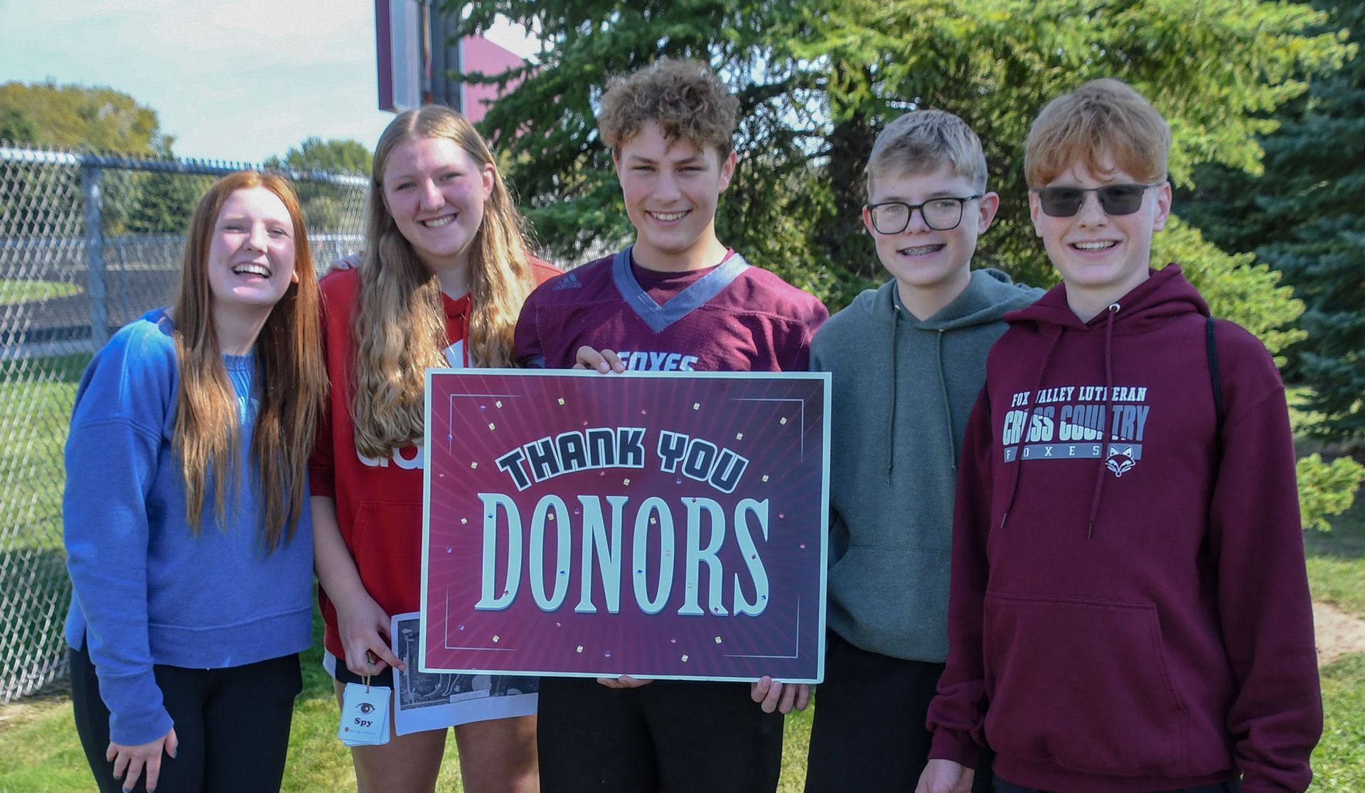 Three smiling boys and two smiling girls standing outside while holding a 