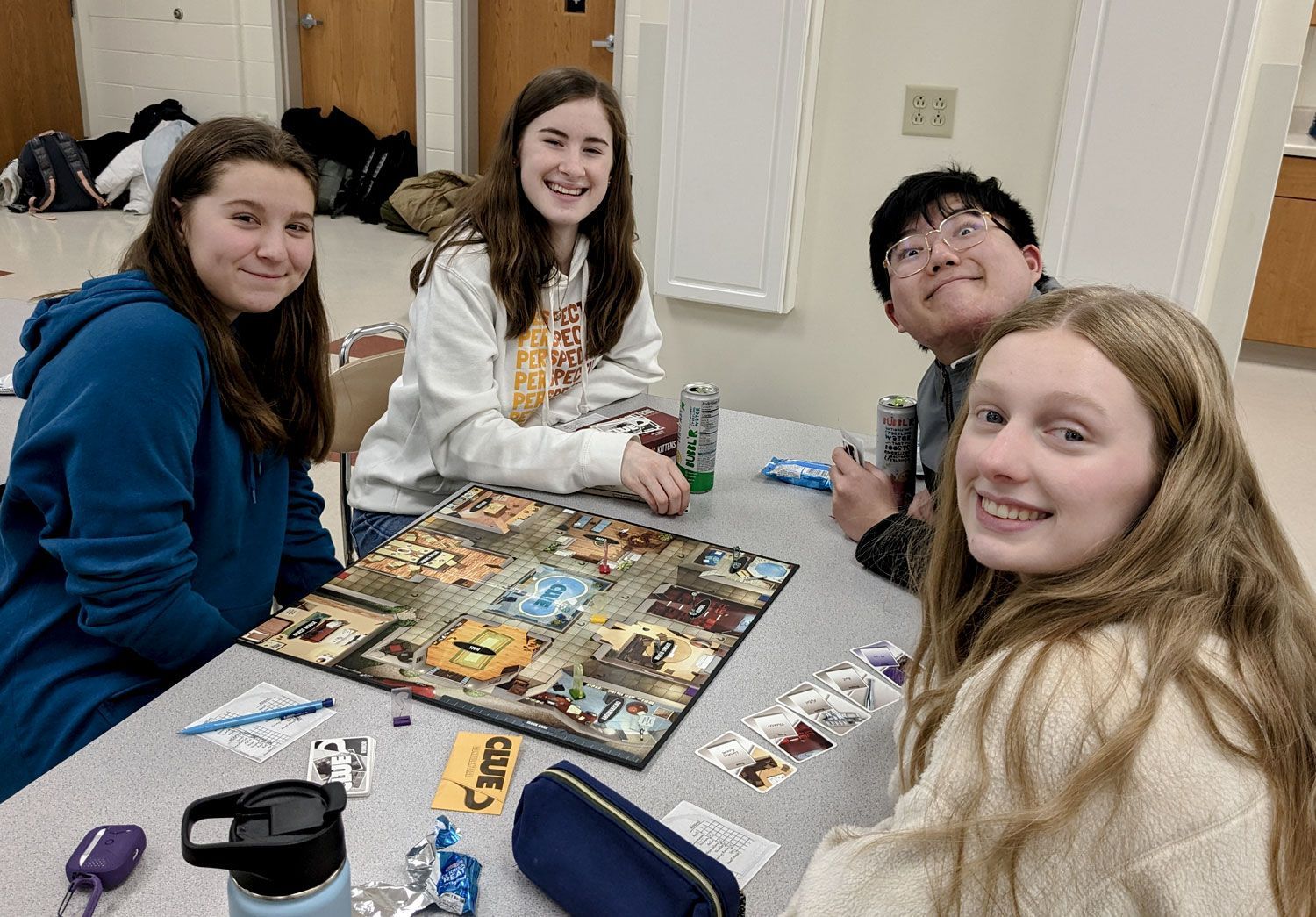 Three girls and one boy smiling at the camera while they sit at a table in a classroom, playing a board game